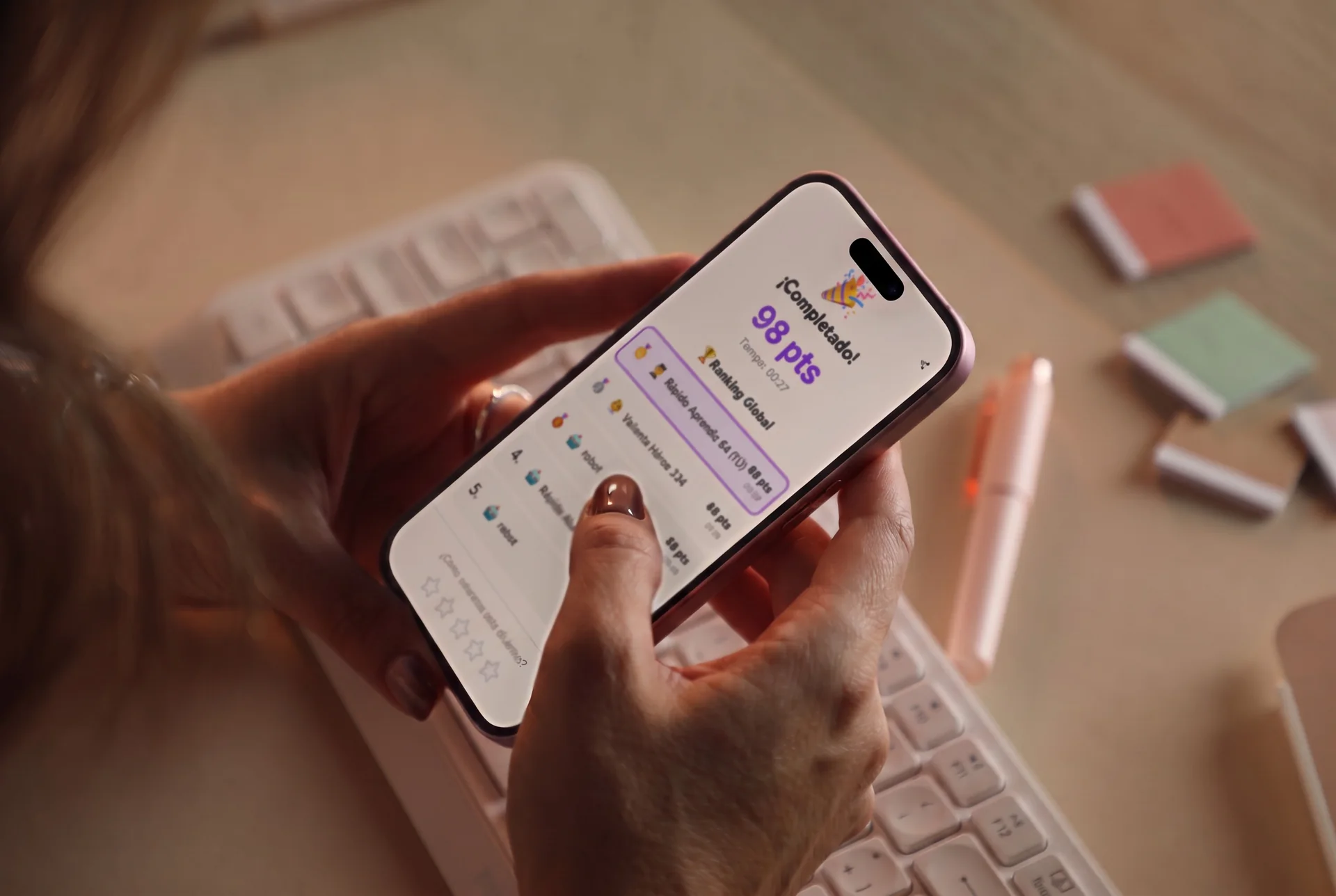 Mano sosteniendo un iPhone con la pantalla de resultado de una dinámica de TaplyLab: ¡Completado! 98 pts y ranking global, sobre un escritorio con teclado y notas adhesivas